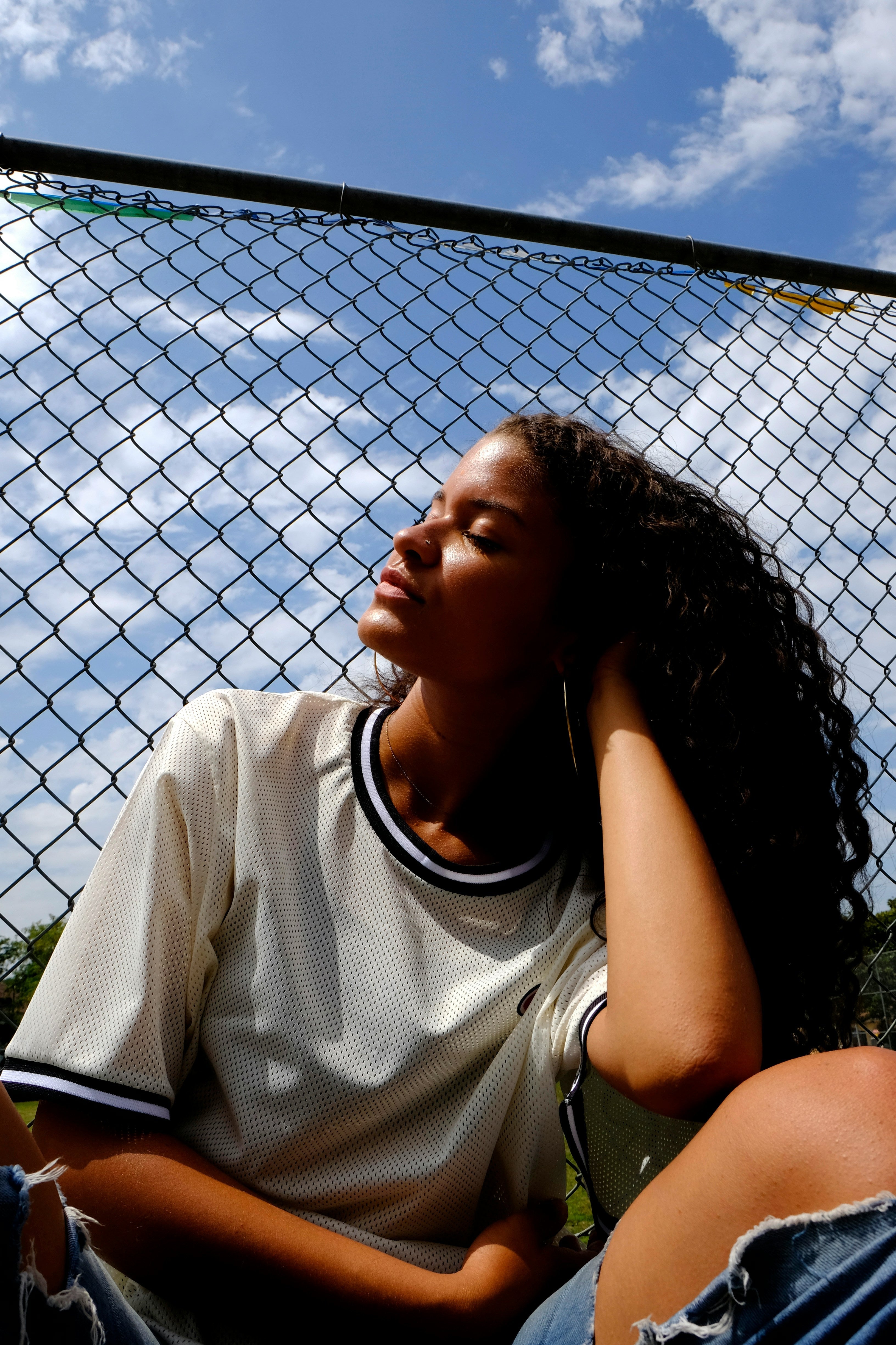 woman outside with blue skys looking up into the sun. She has long curly hair and a athletic shirt. she is sitting in front of a chair link fence